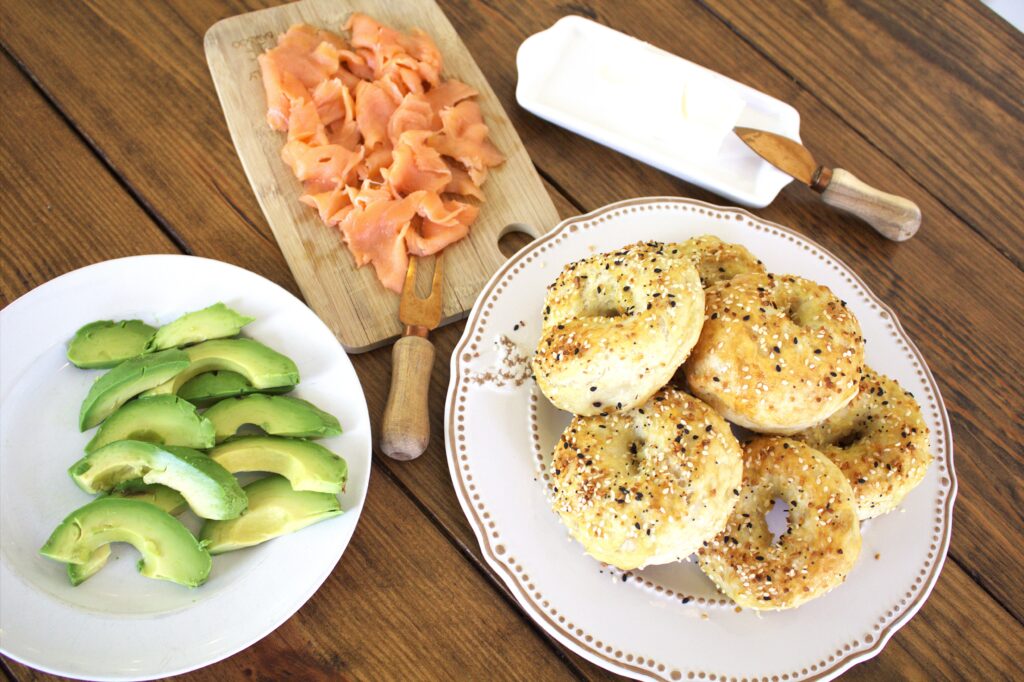 Image of a table holding a plate of sourdough bagels, cream cheese, avocados, and smoked salmon.