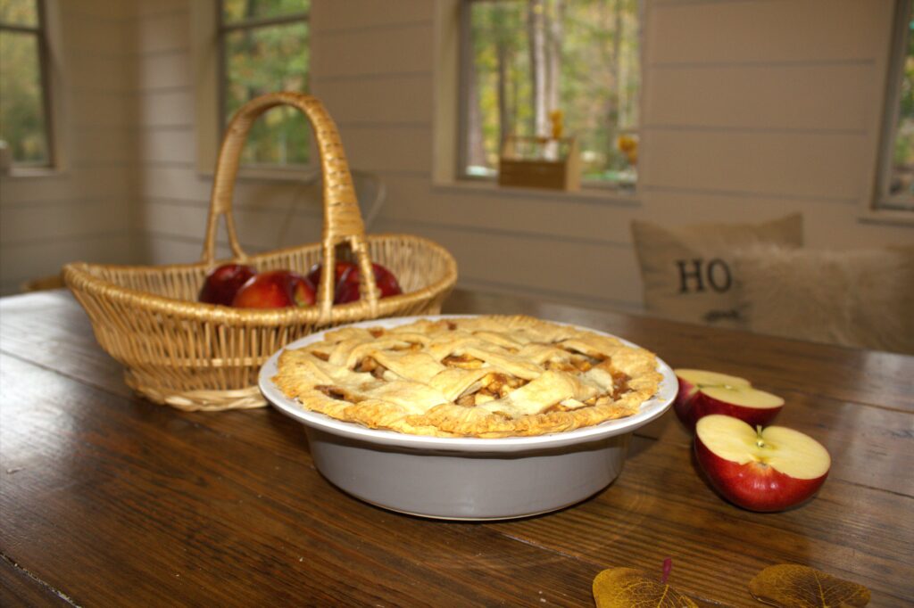 A room with windows and a wood table holding a basket full of apples, an apple pie and a sliced apple