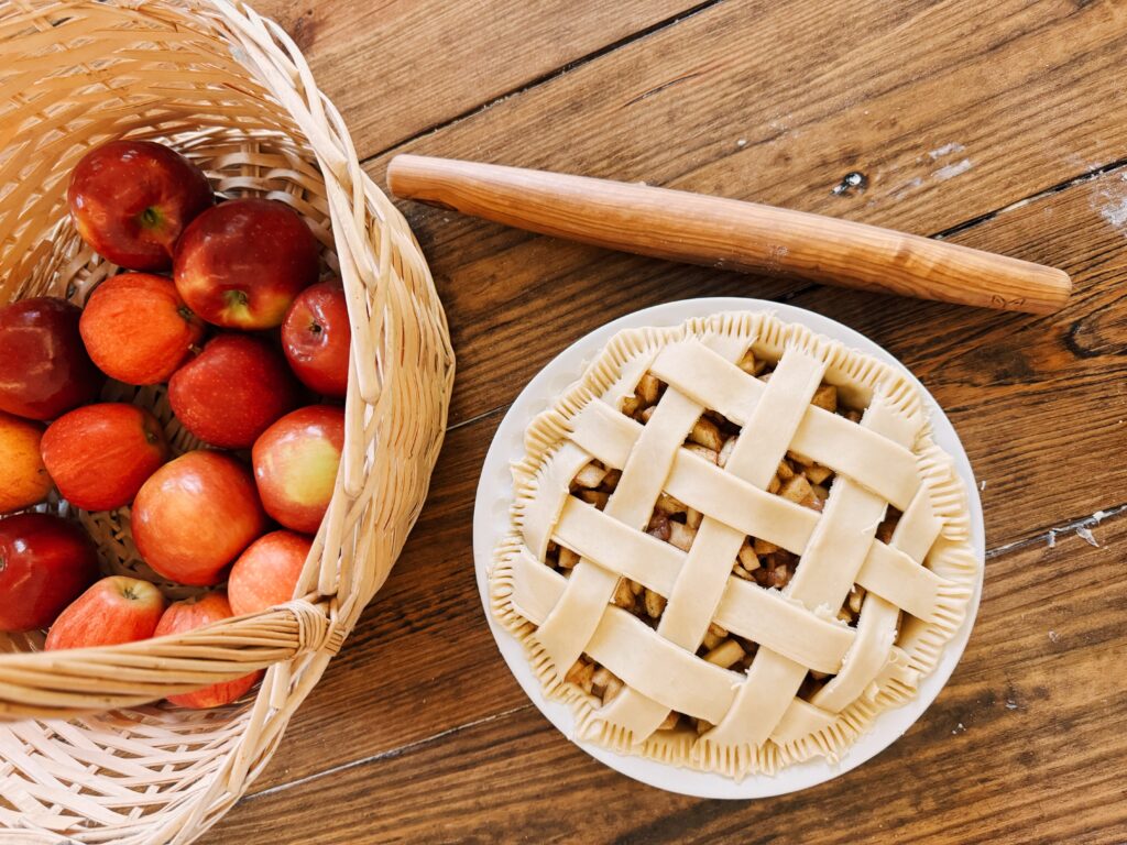 A wood table holding a basket filled with apples, a rolling pin, and and apple pie.