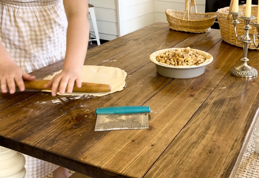 A wooden table holding an apple pie, and a girl using a rolling pin to roll out dough