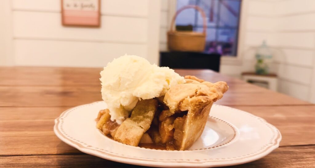 A wooden table holding a white plate with apple pie topped with ice cream