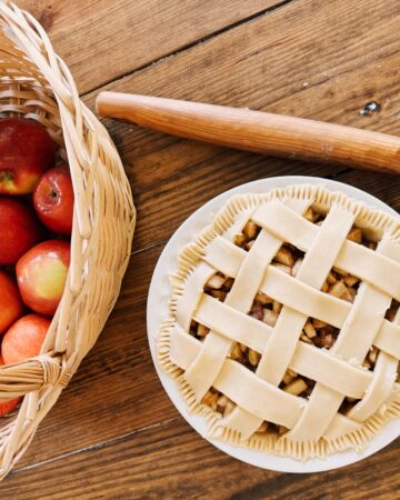 A wooden table holding a rolling pin, a basket of apples, and an apple pie.