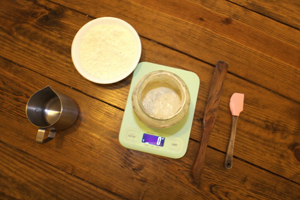 A table with sourdough starter, a stirring stick, flour, water and a scale.