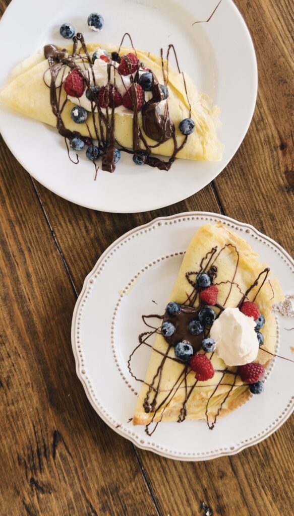 A wood table holding two plates of crepes topped with fruits, chocolate, and whipped cream.