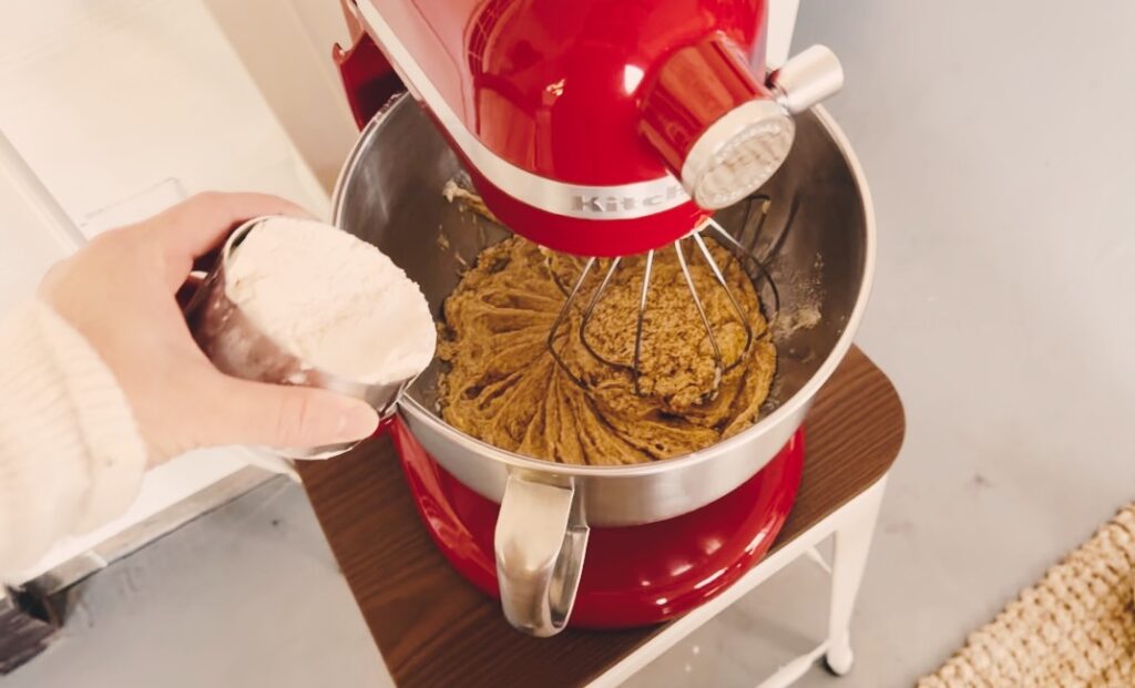 A girl adding flour into a stand mixer containing cookie batter.