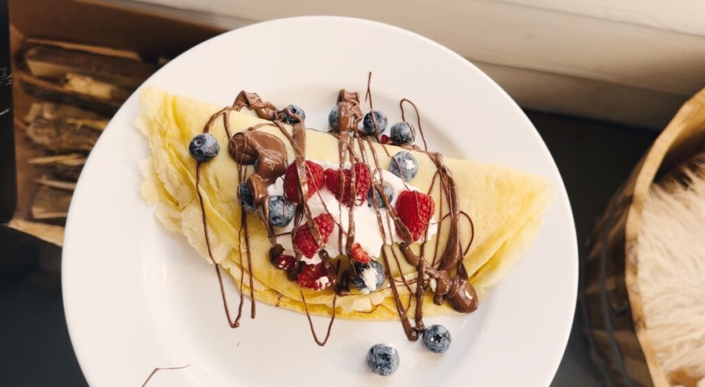 A white plate holding a crepe that is topped with fruits, chocolate, and whipped cream.