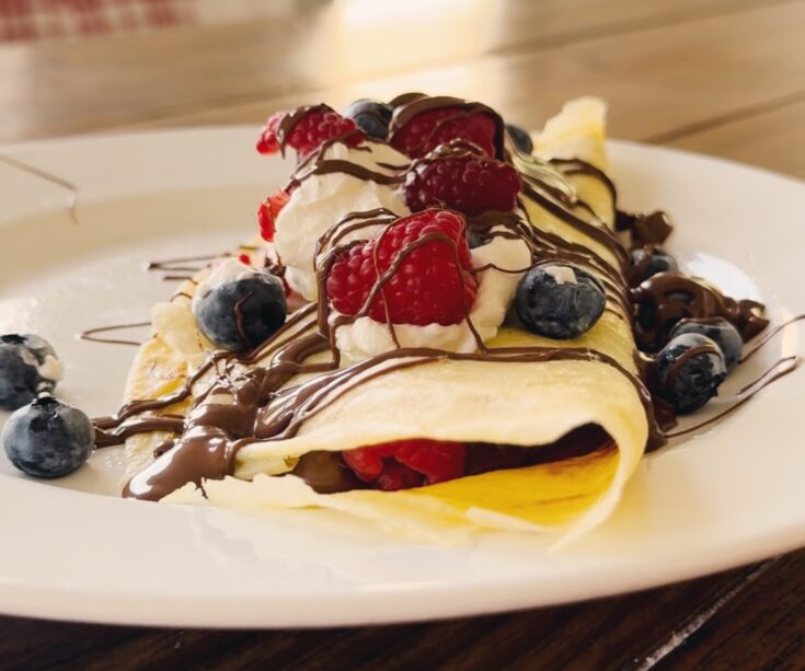 A wood table holding a white plate with a crepe that's topped with berries, chocolate, and whipped cream.