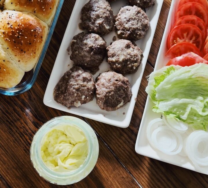 A table spread of fresh vegetables, buns, and burger patties.