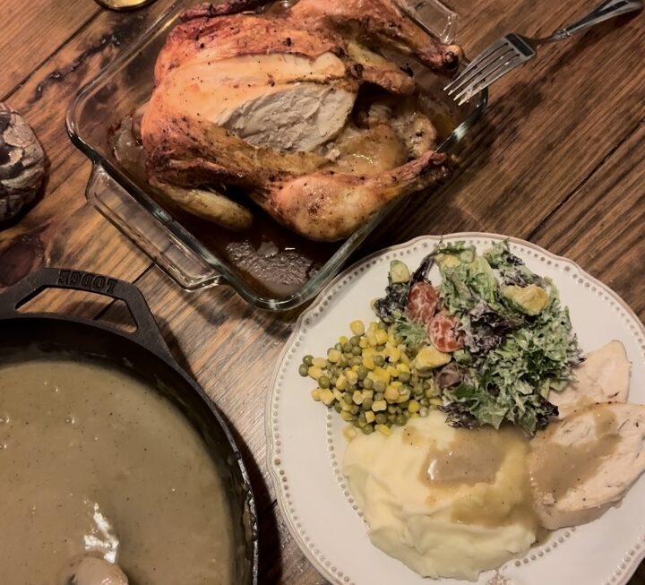 A wood table holding a glass pan with a whole chicken and other dinner foods.